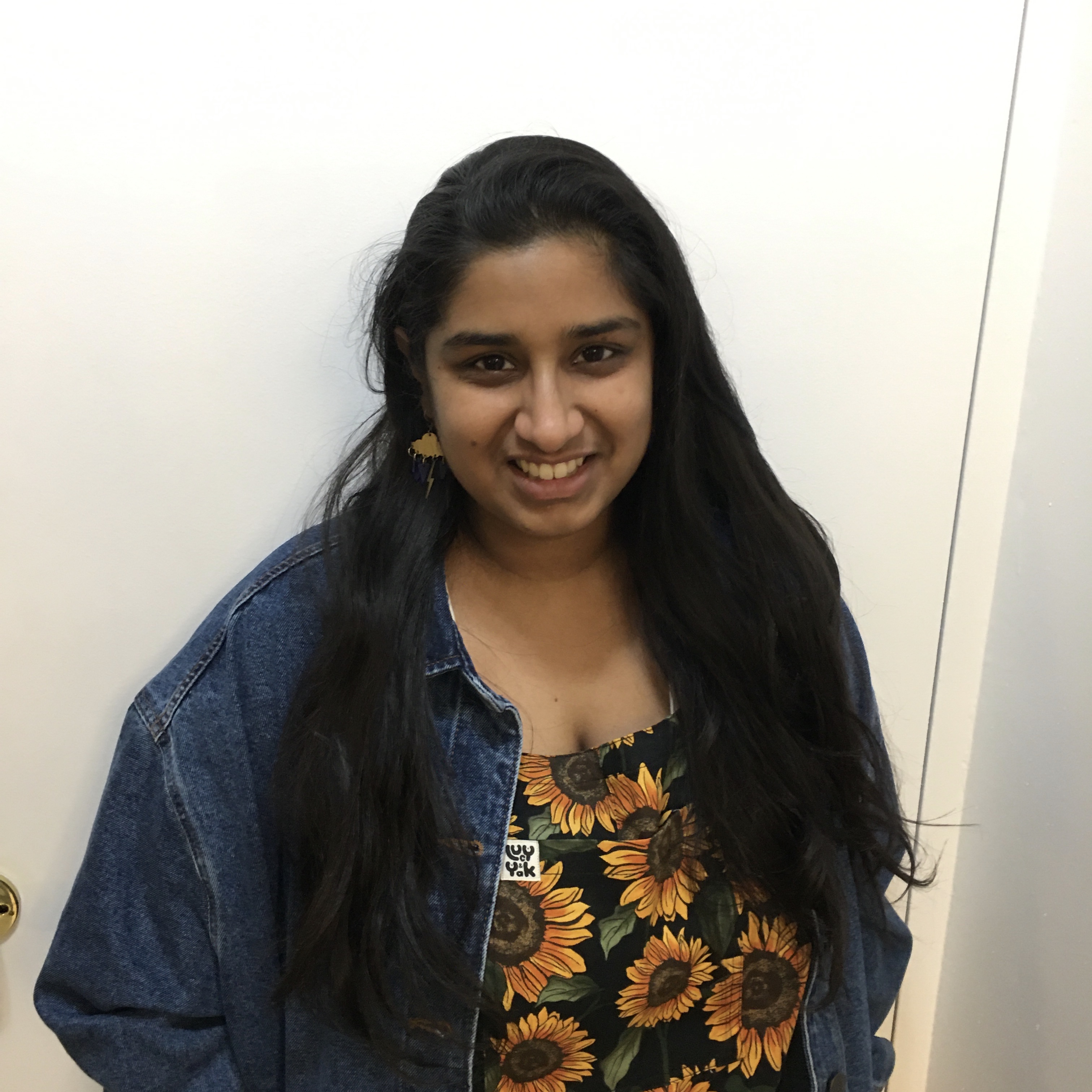 Karuna smiling at the camera wearing a pair of sunflower-patterned overalls and a denim jacket.